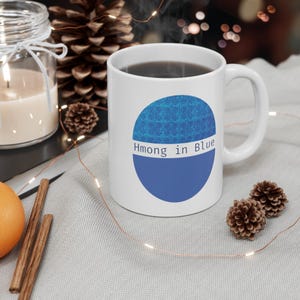 Puede incluir: Taza de cerámica blanca con un diseño azul y el texto "Hmong in Blue". La taza contiene un líquido oscuro y está colocada sobre una superficie gris claro. La escena incluye una vela, piñas y palitos de canela.