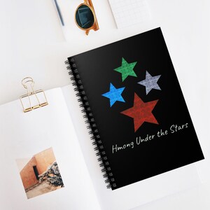 Può includere: Un quaderno nero a spirale con la scritta "Hmong Under the Stars" e quattro stelle colorate. Le stelle sono blu, verdi, grigie e rosse. Il quaderno è aperto su una superficie bianca.