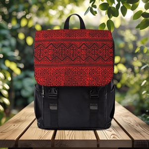 May include: Black backpack with a red flap featuring a geometric pattern in black. The backpack has two side pockets, a top handle, and adjustable straps. The bag is sitting on a wooden table.
