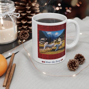 Puede incluir: Taza de cerámica blanca con una imagen escénica de dos perros jugando en un campo con montañas al fondo. La taza tiene un borde rojo y el texto "Wear the Story".