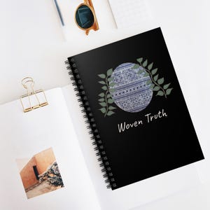 Op de afbeelding: Zwart spiraalgebonden notitieboek met de tekst "Woven Truth" in het wit. De omslag heeft een cirkelvormig ontwerp met blauwe en witte patronen, omlijst door groene bladeren. Het notitieboek is open op een blanco pagina.