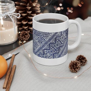 May include: White ceramic mug with a blue geometric pattern. The mug is filled with a dark liquid and has a curved handle. The design features a repeating pattern of squares and lines. The mug is on a light gray surface.