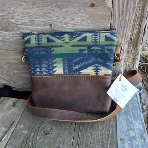 Pode incluir: Uma bolsa transversal com a parte superior estampada e a parte inferior em couro marrom. A parte superior apresenta um design geométrico azul, verde e branco. A bolsa tem uma alça de couro marrom e fecho de zíper.