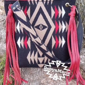 May include: Black and tan geometric pattern crossbody bag with red fringe accents. The bag features a black leather strap and red leather fringe. The bag has a decorative metal tag that says "RobynMarie".