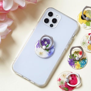 Pode incluir: Cinco suportes para anéis de telefone com um anel prateado e uma base de resina transparente cheia de flores prensadas. As flores são de várias cores, incluindo amarelo, rosa, roxo e azul.