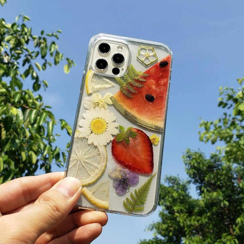 May include: A clear phone case with a pressed flower and fruit design. The design includes a watermelon slice, a strawberry, a lemon slice, and various flowers.