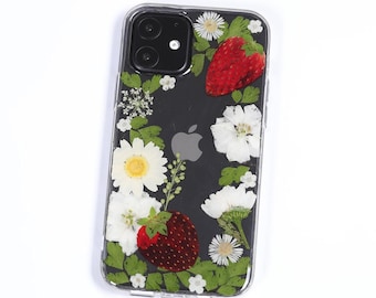 May include: A clear smartphone case featuring a decorative arrangement of pressed dried flowers and two red strawberries. The design includes white daisies, small white blossoms, and green leaves embedded within the transparent material.