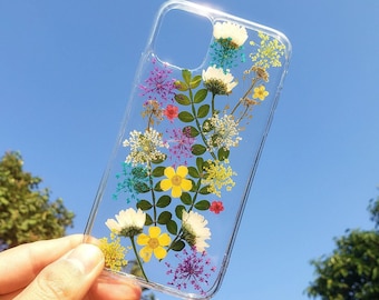 May include: A clear phone case featuring an arrangement of pressed flowers and green leaves. The design includes white daisies, bright yellow buttercups, purple lace flowers, and small yellow clusters, all encased within the transparent plastic.