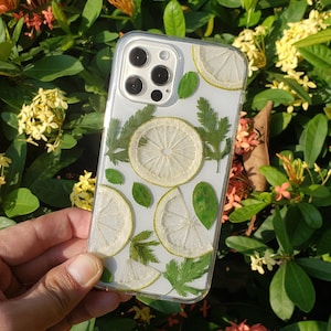 May include: A clear phone case with dried lemon slices and green leaves pressed inside. The case is designed for a smartphone with a triple lens camera.