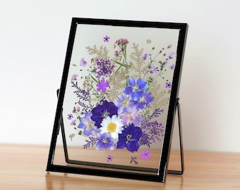 May include: A black framed picture with a pressed flower arrangement. The flowers are shades of purple, blue, and white. The frame has a stand and is sitting on a wooden surface.