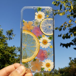 Transparent phone case with natural pressed flowers and citrus slices held up against a clear blue sky, showcasing vibrant lemons and a mix of white daisies and pink florals, capturing the essence of summer, available at SunnyPigStudio.
