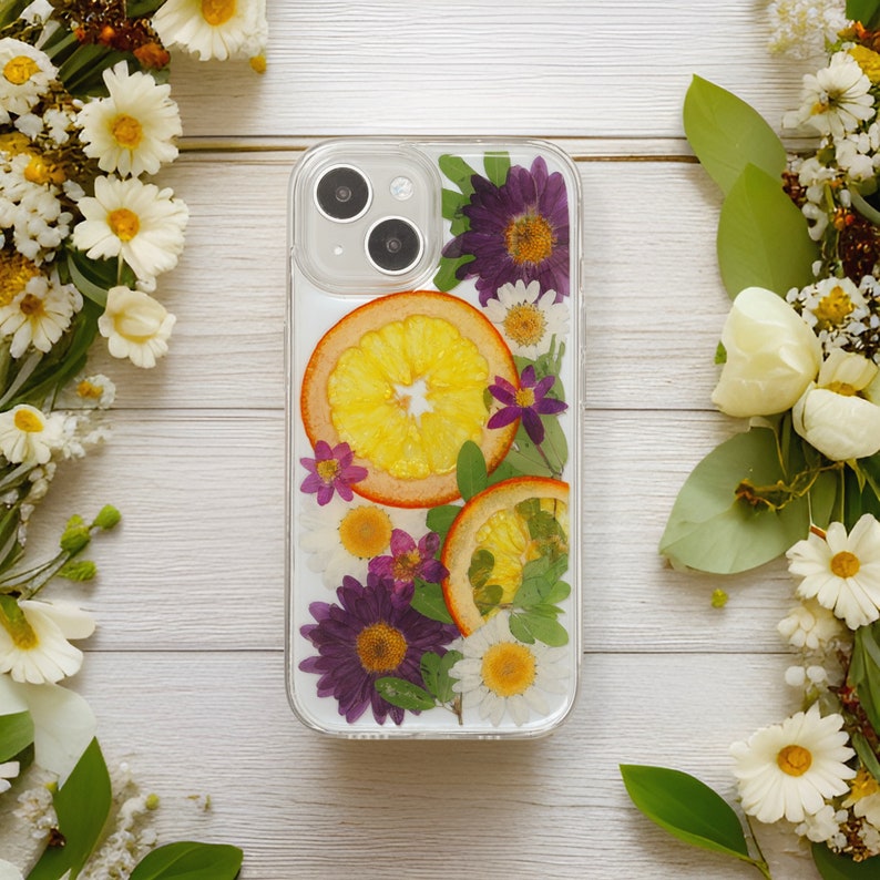 Handcrafted phone case adorned with real pressed flowers and orange slices, displayed on a white wooden background with a bouquet of white and yellow flowers.