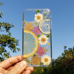 May include: A clear phone case featuring an arrangement of pressed white daisies, small pink and orange flowers, and two translucent citrus slices. The design is embedded within the transparent material, showcasing a botanical and fruit-themed pattern.