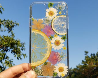Könnte beinhalten: Eine transparente Handyhülle mit einem Arrangement aus gepressten weißen Gänseblümchen, kleinen rosa und orangefarbenen Blüten sowie zwei durchscheinenden Zitrusfruchtscheiben. Das botanische Fruchtmuster ist in das Material eingebettet.