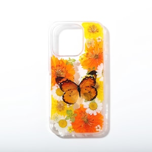 Puede incluir: Una funda de teléfono transparente con una mariposa naranja prensada en el centro, rodeada por un arreglo de flores amarillas y naranjas prensadas y pequeñas margaritas blancas. El diseño está incrustado en el material transparente de la funda.