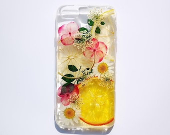 May include: A clear phone case featuring embedded pressed flowers and fruit slices. The design includes pink hydrangea petals, white daisies, small green leaves, and translucent yellow citrus slices arranged across the transparent surface.