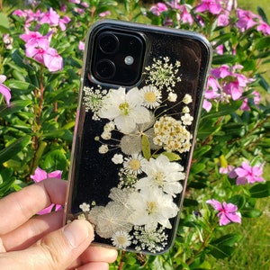 A hand holding a transparent phone case adorned with a variety of delicate white pressed flowers, showcased against a garden with pink periwinkles, highlighting SunnyPigStudio's nature-inspired design.