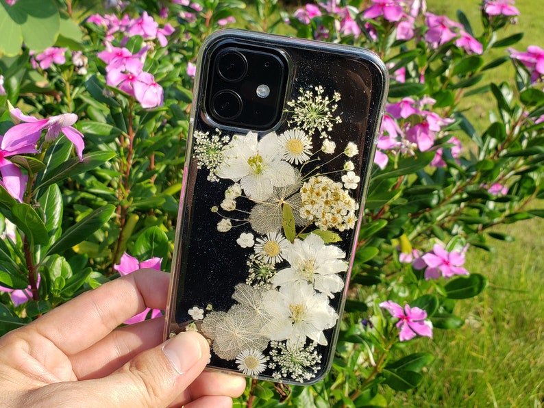 A hand holding a transparent phone case adorned with a variety of delicate white pressed flowers, showcased against a garden with pink periwinkles, highlighting SunnyPigStudio's nature-inspired design.