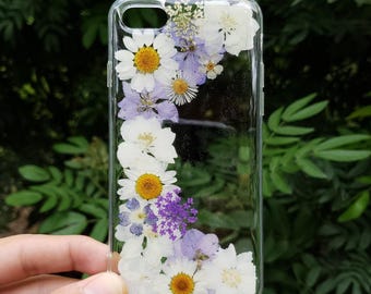 May include: A clear phone case featuring an arrangement of pressed white daisies, purple wildflowers, and small green foliage embedded within the transparent material. The floral design follows the curved edge of the rectangular phone case.
