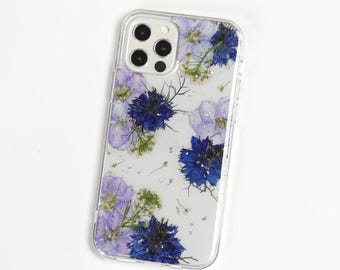 May include: A clear smartphone case featuring an arrangement of pressed blue and light purple flowers embedded within the transparent material. The design includes various floral shapes and green stems, with a cutout for a triple camera system.