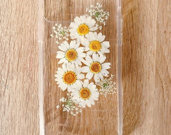 May include: A clear phone case with pressed white daisies and green stems. The flowers are arranged in a cluster in the center of the case.