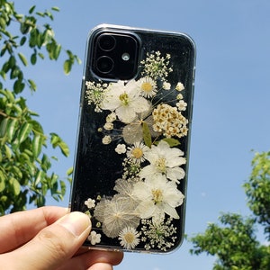 Hand holding a clear phone case with pressed white flowers and sparkles, showcased against a sunny blue sky and green foliage, by SunnyPigStudio, blending nature with everyday tech.