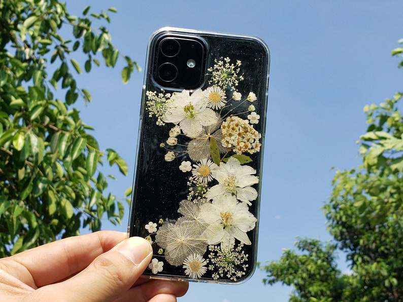 Hand holding a clear phone case with pressed white flowers and sparkles, showcased against a sunny blue sky and green foliage, by SunnyPigStudio, blending nature with everyday tech.