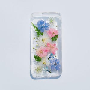 Puede incluir: Una funda transparente para teléfono con flores prensadas en su interior. Las flores son rosas, blancas y azules.