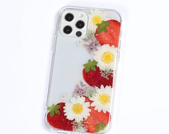 May include: A clear phone case featuring a decorative arrangement of pressed white daisy flowers, red strawberry slices, and small illustrations of cat faces embedded within the transparent material, surrounding the camera lens cutout area.