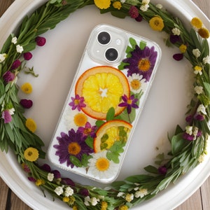 A captivating phone case encircled by a lush wreath, featuring bold purple asters, cheerful daisies, and bright citrus slices, all coming together to form a celebration of floral splendor.
