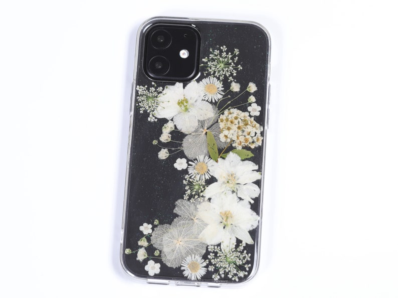 Clear phone case with a touch of glitter, adorned with an arrangement of natural pressed flowers in white and soft beige, accented with green leaves, offering a sparkling and delicate floral design, available from SunnyPigStudio.