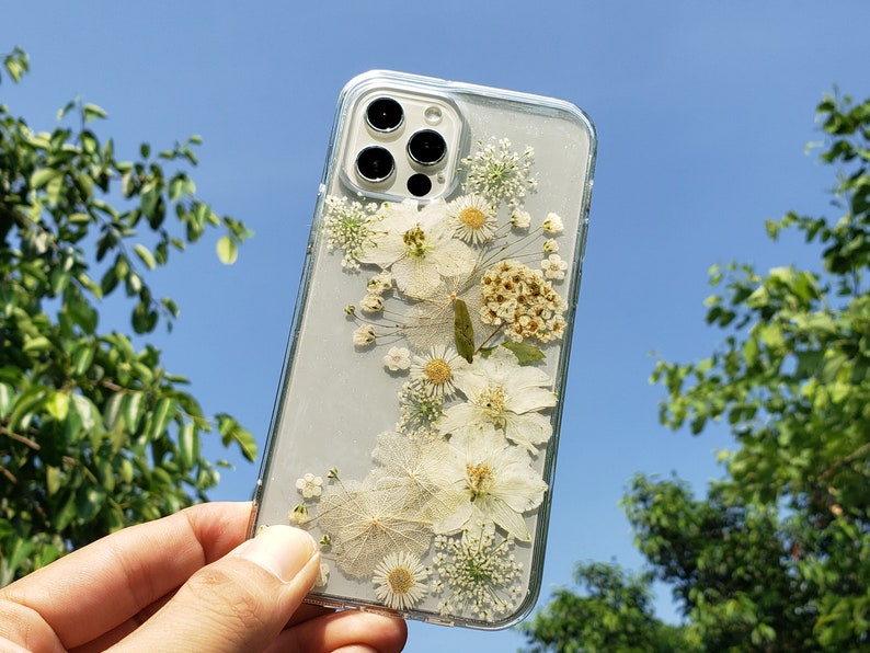 Transparent phone case with white pressed flowers held up to a clear blue sky, capturing the essence of SunnyPigStudio's delicate and natural design aesthetic.