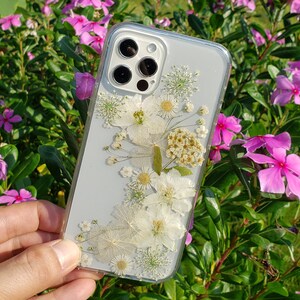 A hand displaying a transparent phone case with a garden-inspired assortment of pressed white flowers, set against a vibrant background of pink periwinkle flowers and lush greenery, crafted by SunnyPigStudio.