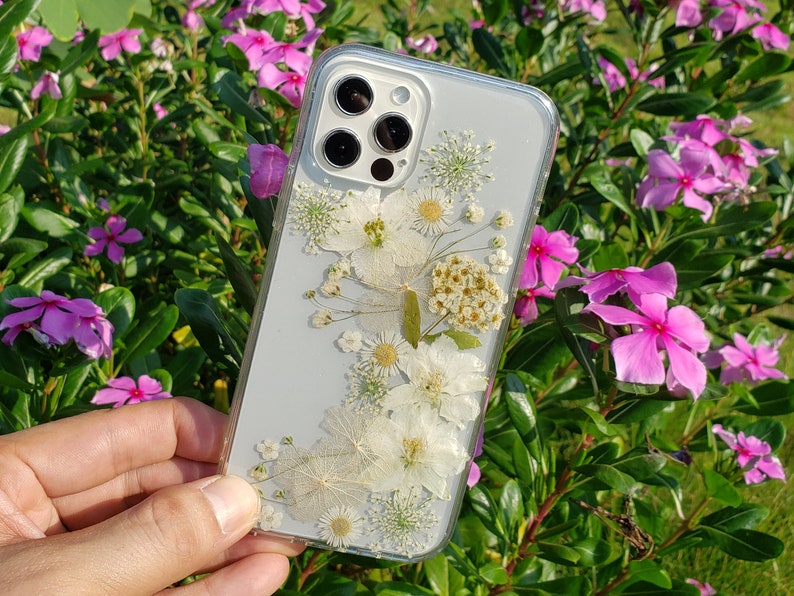 A hand displaying a transparent phone case with a garden-inspired assortment of pressed white flowers, set against a vibrant background of pink periwinkle flowers and lush greenery, crafted by SunnyPigStudio.