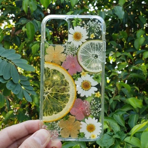 Handheld phone case with a natural botanical theme featuring pressed citrus slices and a variety of flowers including white daisies, set against a lush green foliage backdrop, crafted by SunnyPigStudio.