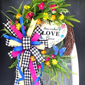 May include: A vibrant, colorful wreath with a heart-shaped sign that reads "Home is where LOVE Grows." The wreath features a variety of flowers, greenery, and a large bow with black and white checkered ribbon, pink, yellow, and blue ribbons.