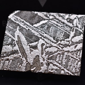 May include: Close-up of a square-cut, metallic meteorite. The surface displays a unique Widmanstätten pattern, with intersecting bands of light and dark gray. The intricate design is set against a black background, highlighting the meteorite's texture.