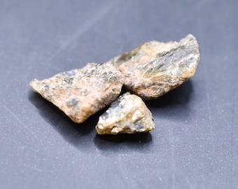 May include: Three pieces of rough, gray and brown, natural, unpolished, mineral rock. The rocks are on a dark gray surface.