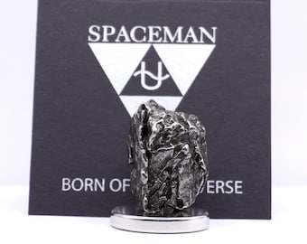 May include: A dark, textured meteorite on a silver base, with a black background card. The card features the word "SPACEMAN" above a white triangle logo and the text "BORN OF UNIVERSE".