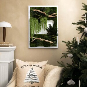 May include: A framed wall art piece featuring a vibrant green moss design with natural wood branches and cascading greenery. A pillow with a winter tree graphic and the text "Winter Woods" rests on a chair. A Christmas tree with ornaments is in the background.