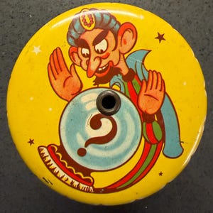 May include: A vintage, yellow fortune-telling toy featuring a cartoon genie with a blue turban and cape. The genie is gesturing towards a blue crystal ball with a question mark. The toy has a hole in the center and is decorated with stars.