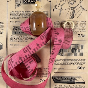 May include: A vintage novelty toy set featuring a pink measuring tape, a small brown container with a white top, and text from an advertisement. The text reads "Joke Novelties".