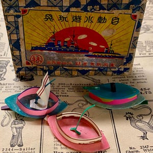 May include: Vintage toy boats in pink, blue, and teal with white sails and green masts. The boats are displayed with a box featuring a ship illustration and Japanese text. The box has a blue and yellow design.
