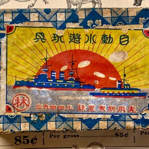 May include: Vintage Japanese box with a nautical theme. The box features a vibrant illustration of a red sun setting over blue ships, with Japanese text. The box has a blue and white patterned border and shows signs of age.