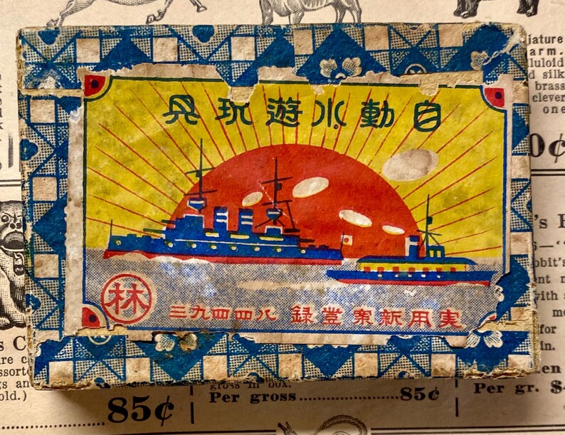 May include: Vintage Japanese box with a nautical theme. The box features a vibrant illustration of a red sun setting over blue ships, with Japanese text. The box has a blue and white patterned border and shows signs of age.