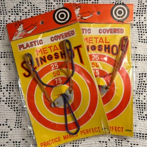 May include: Two plastic-covered metal slingshots in original yellow and orange packaging. Each package features a target graphic with point values 25, 50, and 75, an illustration of a person using a slingshot, and the text "PLASTIC COVERED METAL SLINGSHOT" and "PRACTICE MAKES PERFECT".