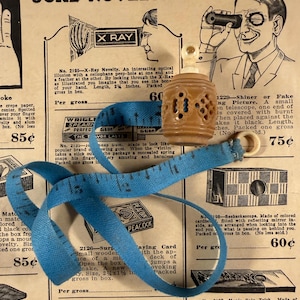 May include: An antique novelty advertisement featuring a blue measuring tape, a carved wooden object, and illustrations. The text includes "JOKE NOVELTIES" and descriptions of various novelty items.