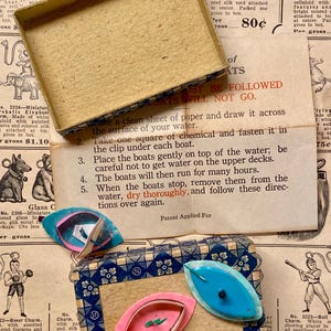 May include: Vintage toy boat charms in a decorative box. The charms are in shades of pink and blue, with a leaf-like shape. The box has a blue and white patterned design. Instructions for use are visible.