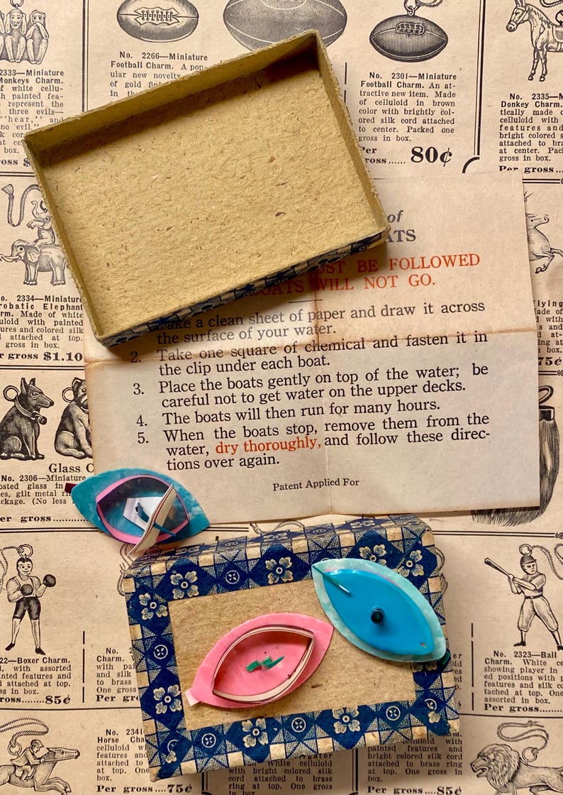 May include: Vintage toy boat charms in a decorative box. The charms are in shades of pink and blue, with a leaf-like shape. The box has a blue and white patterned design. Instructions for use are visible.