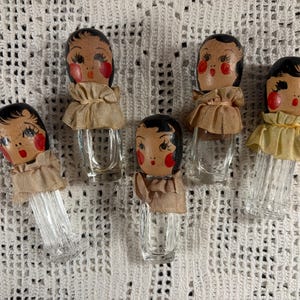 May include: Five clear glass bottles with fluted or smooth bodies, each topped with a hand-painted wooden head featuring black hair, red cheeks, and red lips. Each bottle is adorned with a gathered beige fabric collar tied around the neck.